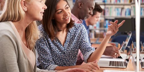 Een groep van vier mensen zit aan een houten tafel. Twee hiervan staan op de voorgrond, ze zijn bezig op een computer en voeren een gesprek. De andere twee personen zijn in de achtergrond bezig op hun laptop.