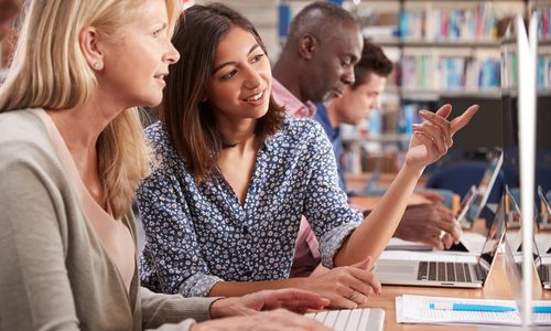 Een groep van vier mensen zit aan een houten tafel. Twee hiervan staan op de voorgrond, ze zijn bezig op een computer en voeren een gesprek. De andere twee personen zijn in de achtergrond bezig op hun laptop.