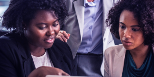 Close-up van drie mensen bij een laptop, één van hun wijst naar het scherm terwijl een ander meekijkt. De achterste persoon is alleen van hun nek tot middel te zien