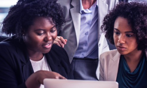 Close-up van drie mensen bij een laptop, één van hun wijst naar het scherm terwijl een ander meekijkt. De achterste persoon is alleen van hun nek tot middel te zien